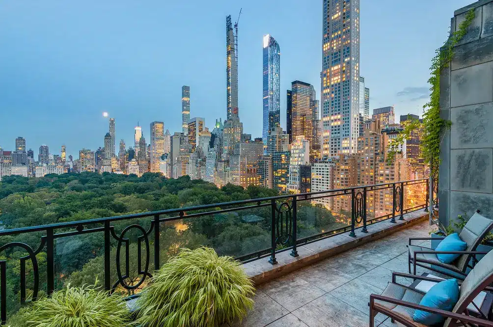 Balcony terrace with outdoor chairs and blue cushions overlooking a dense Manhattan skyline and a park below at dusk, with potted plants in the foreground.