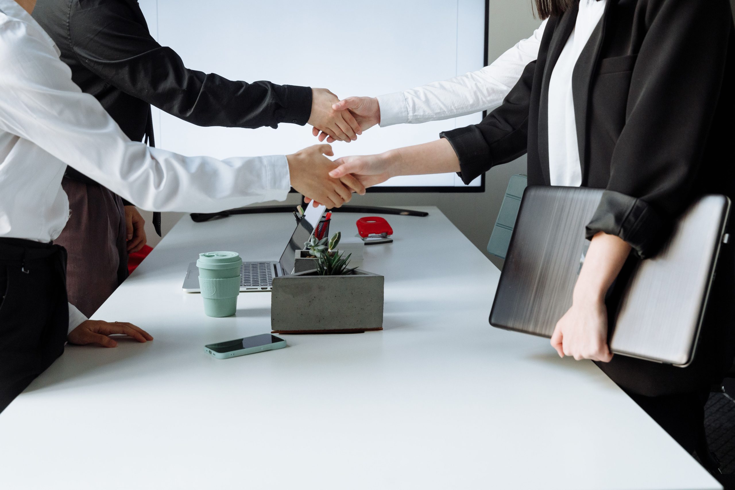 Two professionals shake hands across a white office desk, with laptops and office supplies nearby.